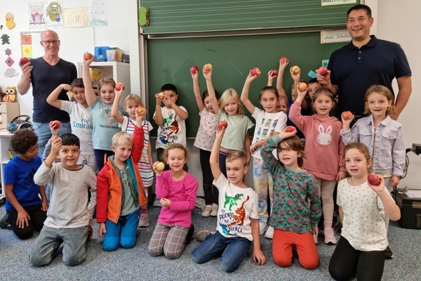 18 Kinder und zwei Erwachsene als Gruppe in einem Klassenzimmer. Jeder hlt einen Apfel in der Hand. 